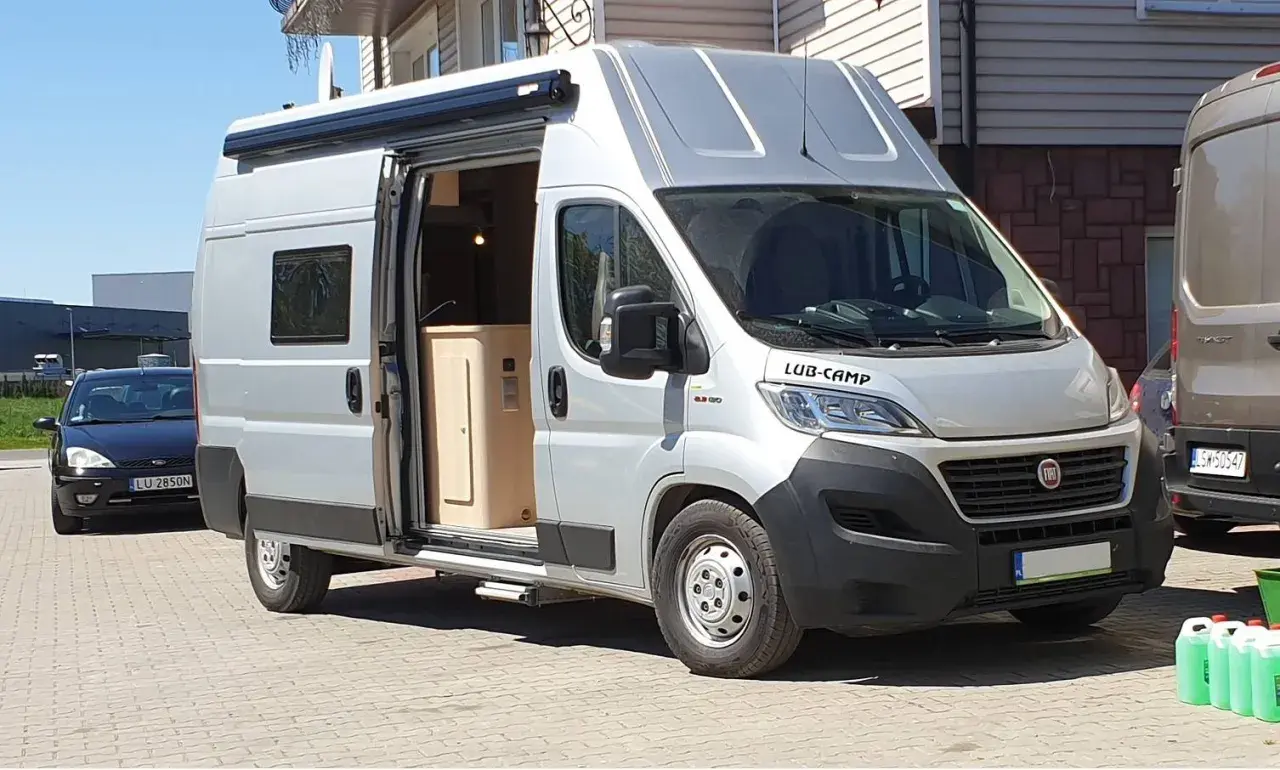 Srebrny kamper Ducato L3H3, z otwartymi drzwiami, ukazuje wnętrze. Idealny do podróży, jego wymiary pozwalają na komfort.