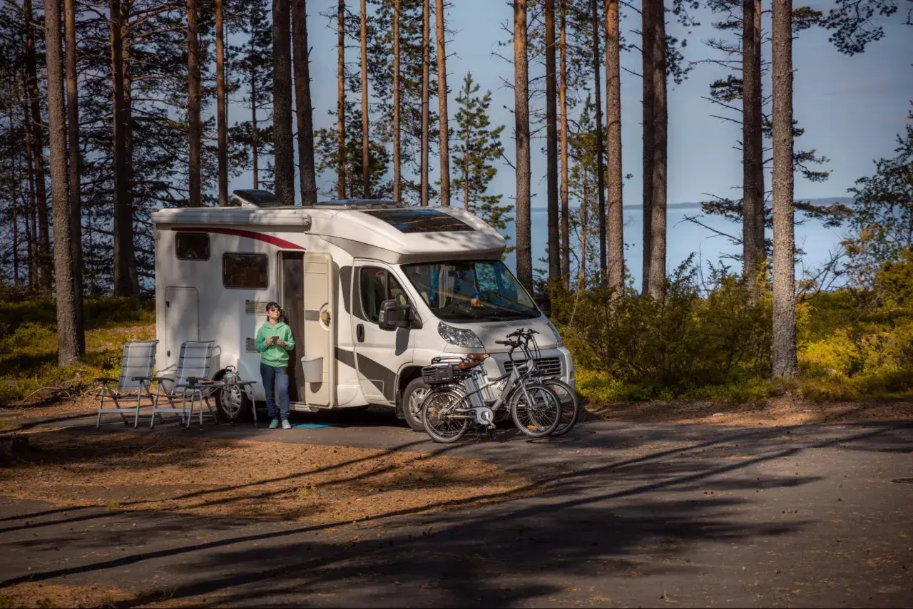 Kamper zaparkowany w lesie, obok jeziora. Na pierwszym planie dwa rowery, obok kampera siedzi osoba. Miejsce idealne na wypoczynek, pogoria 4 parking.