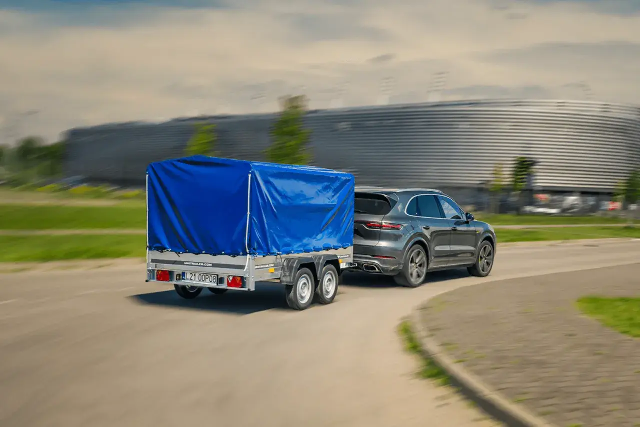 Szare Porsche Cayenne z niebieską plandeką na przyczepie dwuosiowej. Czy można ciągnąć przyczepę dwuosiową na kat. B?