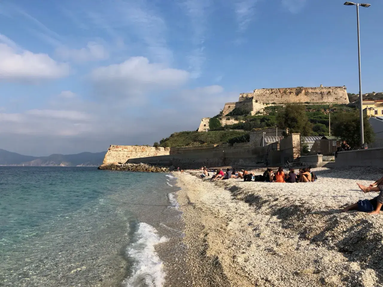 Plaża na wyspie Elba we Włoszech, z turkusową wodą i kamienistym brzegiem. W tle widać zabytkową fortecę na wzgórzu.