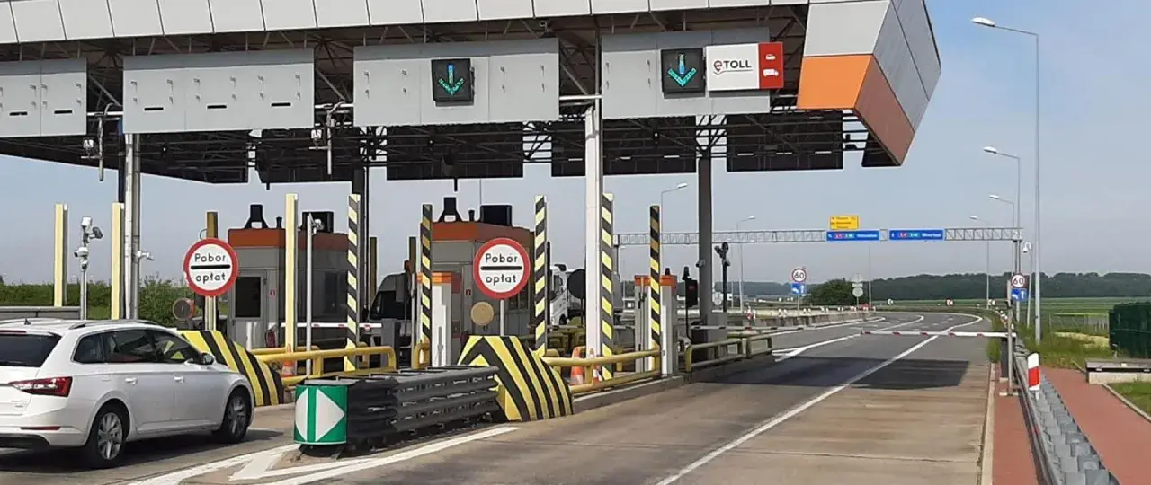 Biały samochód z przyczepą zbliża się do punktu poboru opłat za przejazd autostradą. Widać bramki i znaki informujące o opłatach.