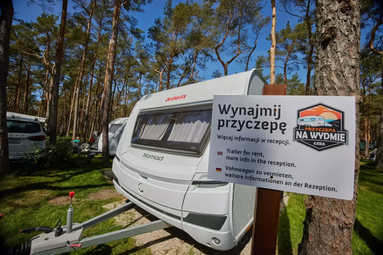 Biała przyczepa kempingowa nad morzem, otoczona sosnowym lasem. Tablica informuje o możliwości wynajmu.