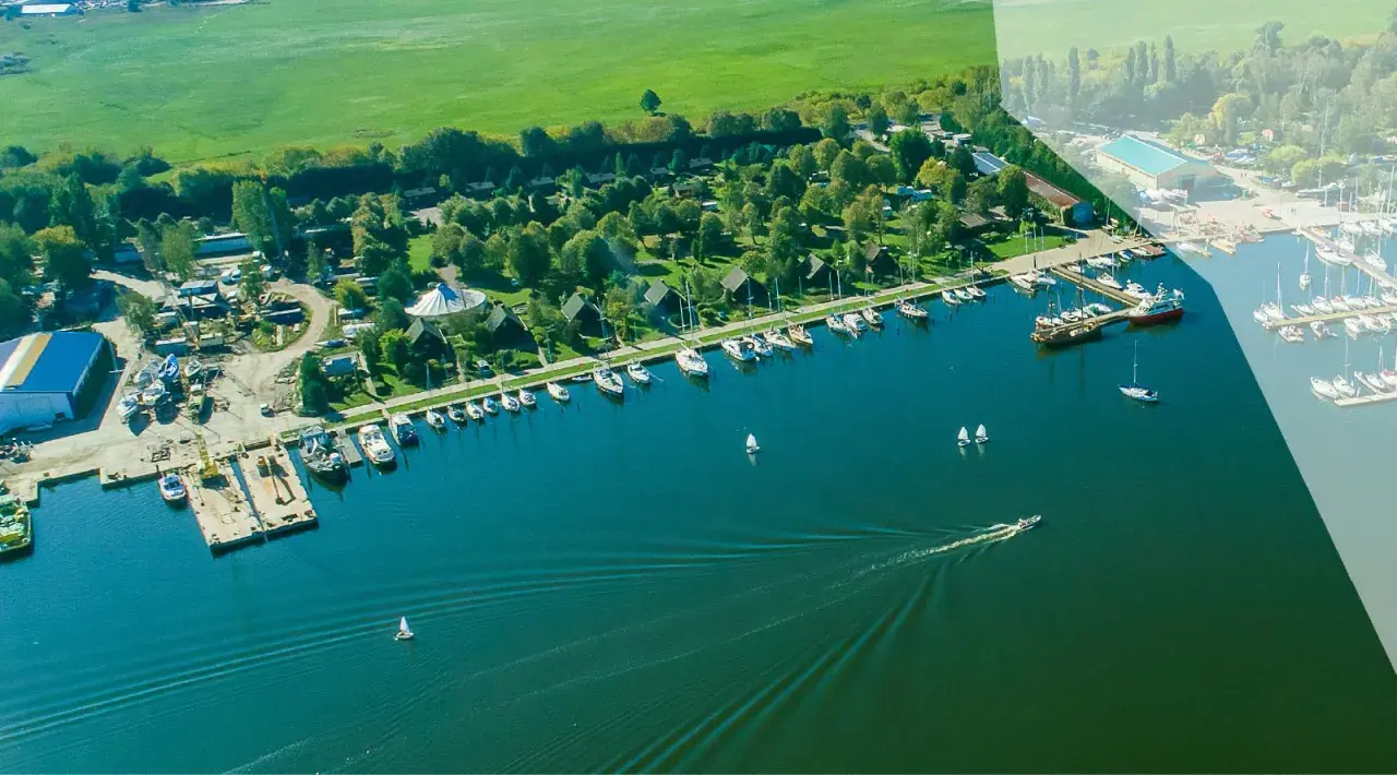 Widok z lotu ptaka na camping marina PTTK z licznymi łodziami i żaglówkami zacumowanymi przy brzegu, otoczony zielenią i polami.