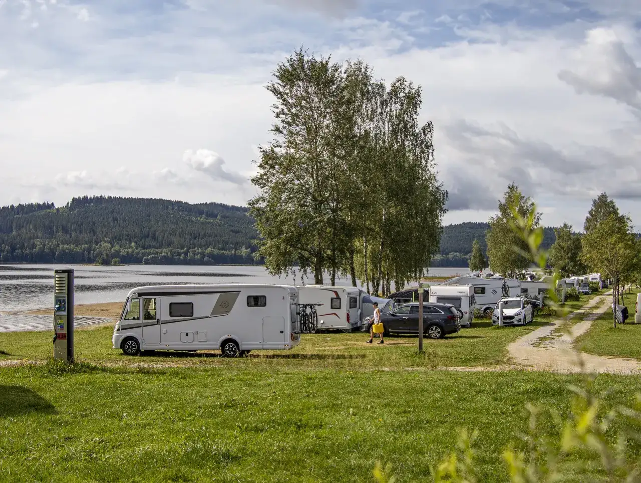 Kampery i przyczepy kempingowe na campingu Lipno nad jeziorem, otoczone zielenią i drzewami.