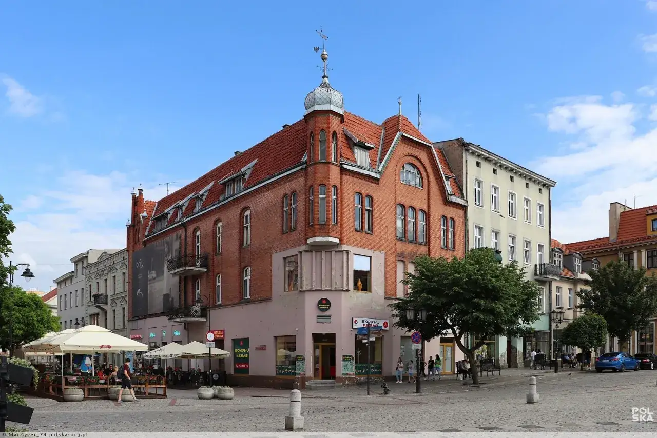 Tętniący życiem rynek w Gnieźnie, z kamienicami, kawiarnianymi ogr&oacute;dkami i spacerującymi ludźmi.