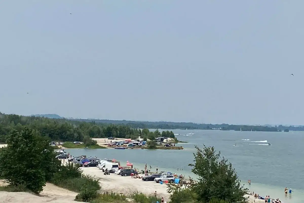 Plaża nad jeziorem, mn&oacute;stwo samochod&oacute;w i namiot&oacute;w tworzy parking przy Pogorii 4. Ludzie odpoczywają, pływają i jeżdżą na skuterach wodnych.