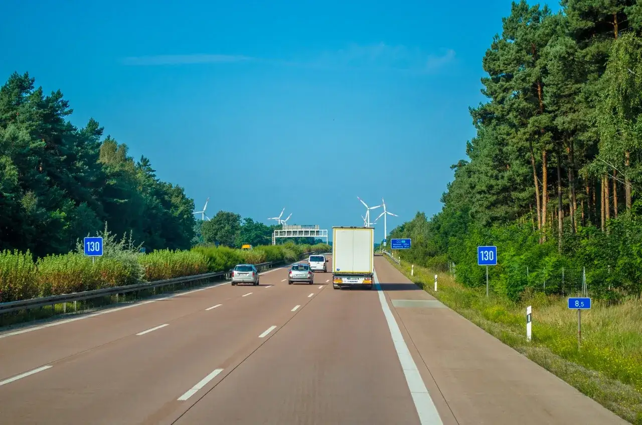 Niemieckie autostrady płatne dla obcokrajowc&oacute;w. Samochody i ciężar&oacute;wka jadące po pustej autostradzie, otoczonej drzewami i polami wiatrak&oacute;w.