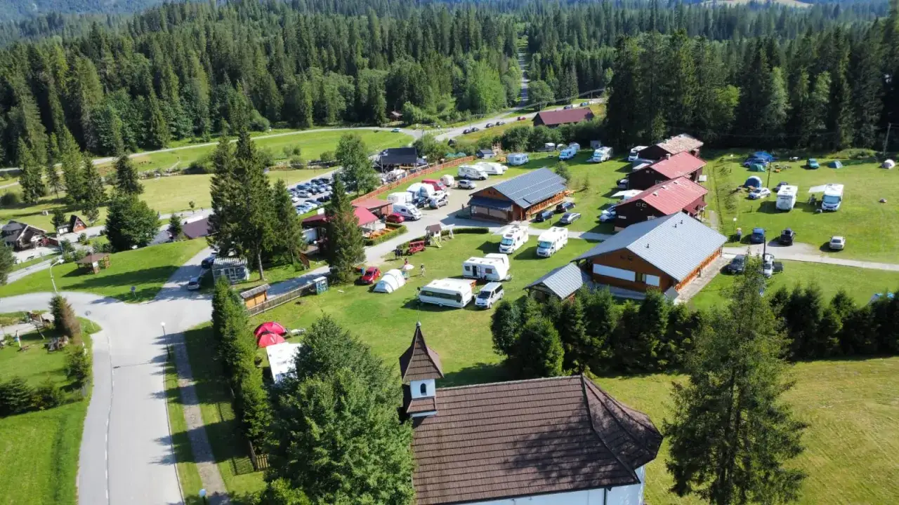 Widok z lotu ptaka na kemping Oravice. Widać tu wiele kamper&oacute;w, przyczep kempingowych, namioty i budynki mieszkalne, otoczone zielenią i lasem.