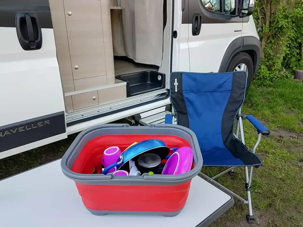 Gotowe naczynia do zmywania na kemping. Składana miska z akcesoriami i niebieskie krzesło obok kampera.