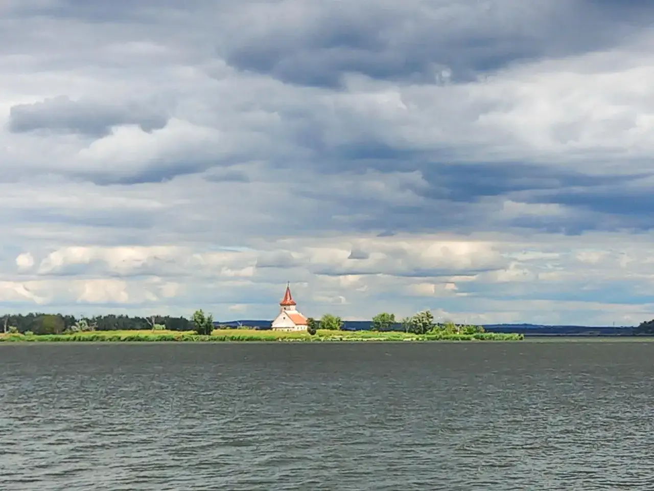 Kościółek na wyspie nad jeziorem w Mikulovie, otoczony zielenią, pod burzliwym niebem.