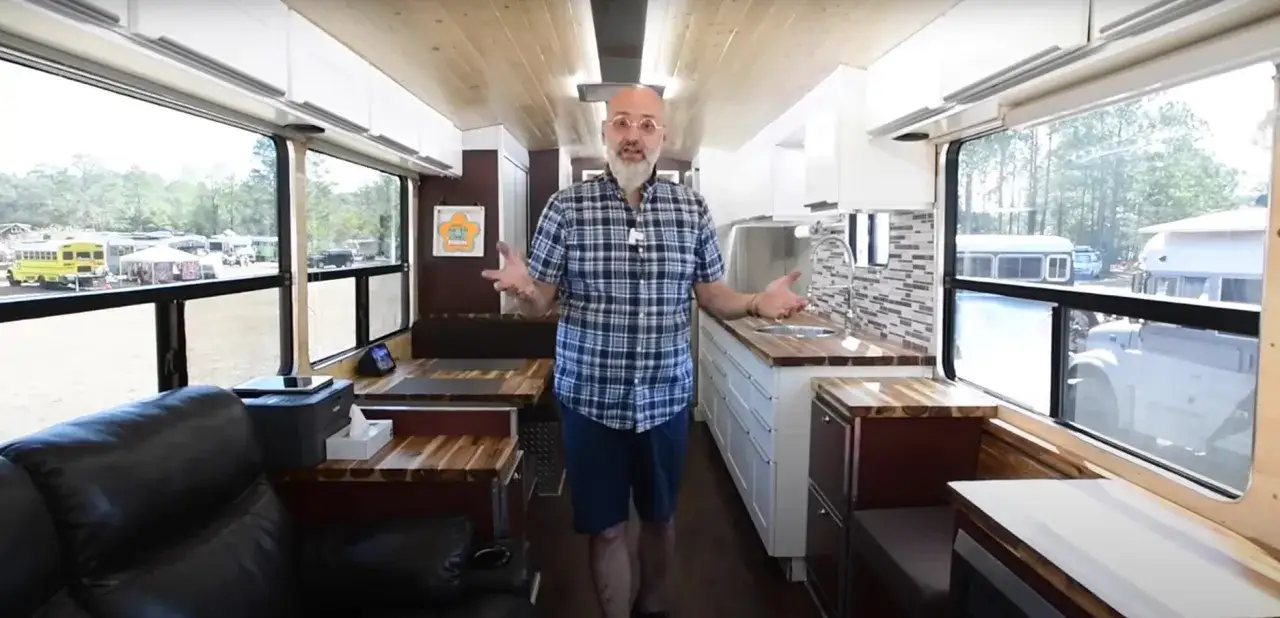 Mężczyzna prezentuje wnętrze swojego domu z autobusu, pokazując kuchnię i miejsce do siedzenia. Widać też inne autobusy na zewnątrz.