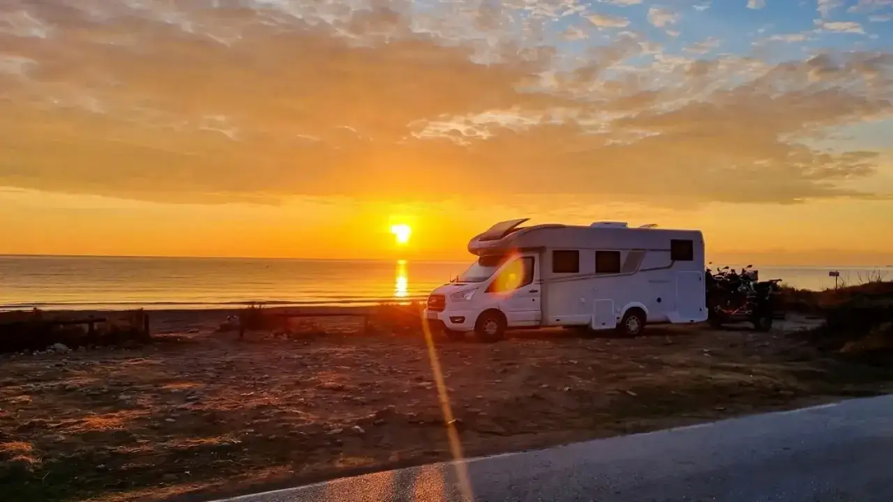 Kamperem nad morzem na dziko. Zachód słońca maluje niebo na pomarańczowo, a pojazd stoi na plaży, gotowy na nocleg.