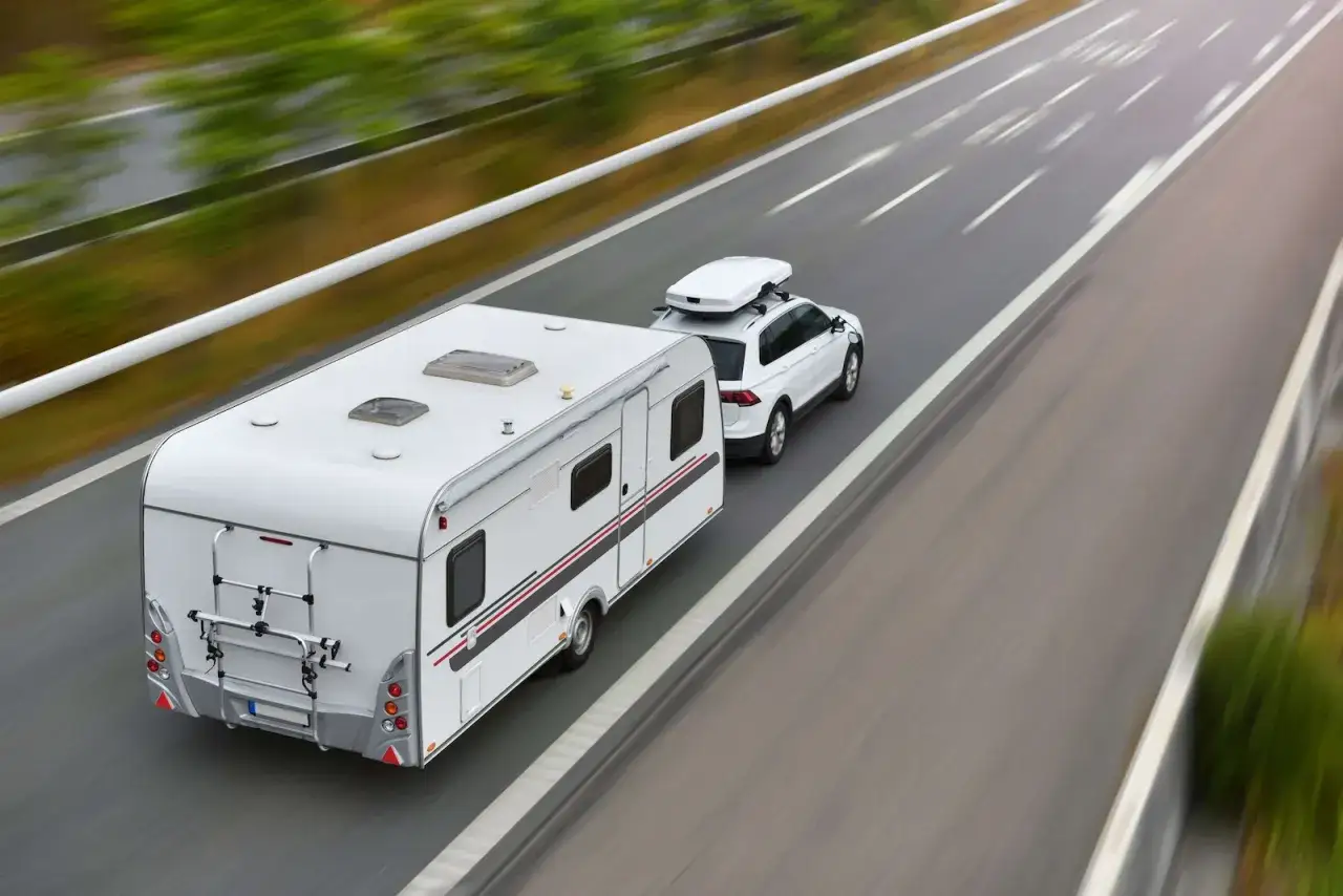 Biały samoch&oacute;d z przyczepą kempingową i boxem dachowym jedzie autostradą. Płatności za przejazd autostradą z przyczepą mogą być wyższe.